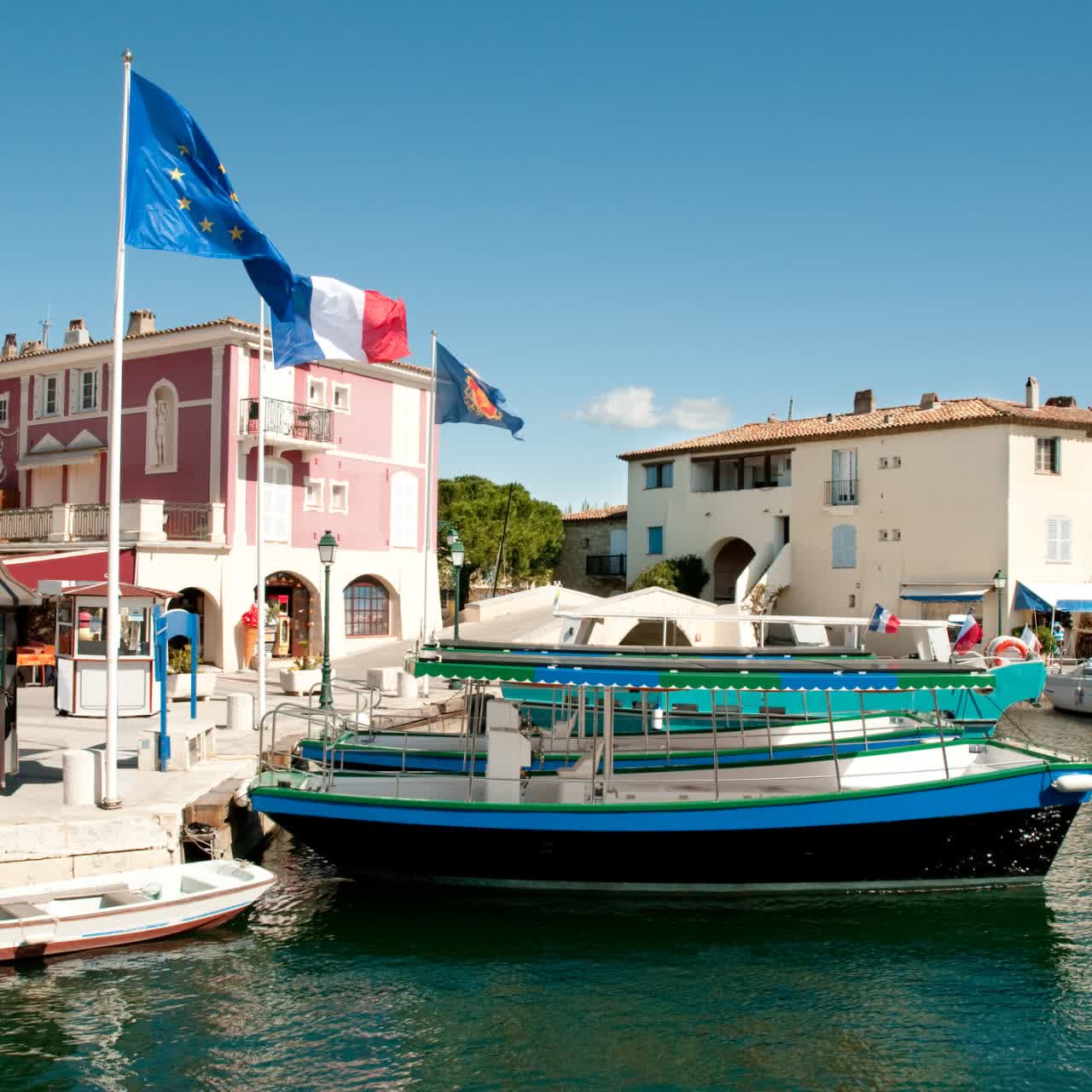 Quai de Port Grimaud avec drapeaux européens et français, maisons colorées et bateaux amarrés